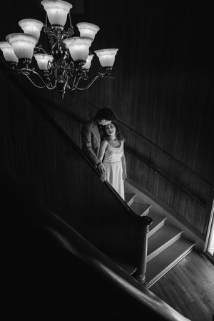 Black and white photo of a couple standing on a staircase beneath an elegant chandelier in a dimly lit interior.