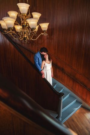 Bride and groom pose on elegant staircase with gold chandelier and wood-paneled walls in romantic wedding portrait.