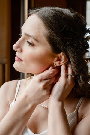 Bride putting on pearl earrings while wearing a white dress and bracelet, preparing for her wedding day.