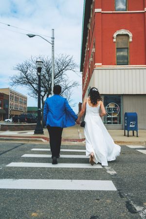 Newlywed couple crossing a zebra crossing, bride in white gown and groom in blue suit, holding hands in urban street.