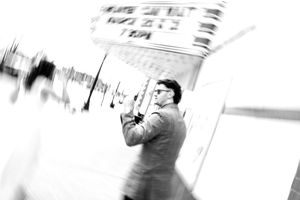 Black and white photo of a man in a suit holding a camera outdoors with motion blur effect.