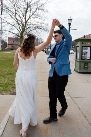 A bride and groom dancing joyfully on a sidewalk, the groom in a blue blazer spinning his bride in a white dress.