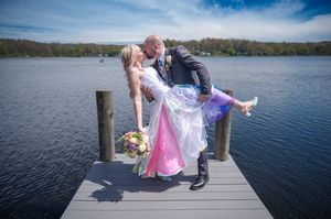 Bride and groom share a romantic dip kiss on a lakeside dock, bride holding colorful bouquet in wedding attire.