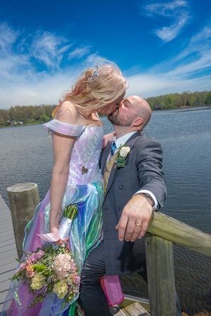 Bride and groom sharing a kiss on a lakeside dock, bride in colorful gown holding bouquet, sunny blue sky backdrop.