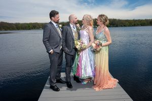 Two couples in formal wedding attire pose on a dock overlooking a serene lake under a bright blue sky.