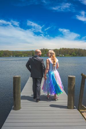 Couple in formal wear holding hands on a lakeside dock under a blue sky with wispy clouds and forested hills.