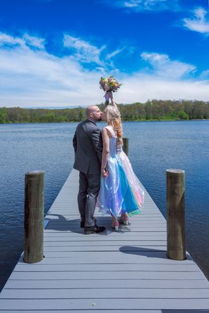 Bride and groom kissing on a lakeside dock, bride holding colorful bouquet aloft in a rainbow gown under blue skies.