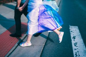 Bride in colorful gown and rainbow sneakers jumping on a crosswalk with groom in black pants.