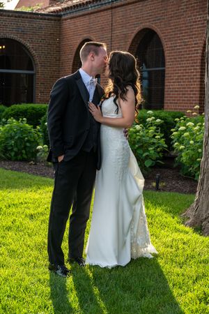 Bride and groom share a romantic kiss on lush green lawn outside brick building with arched windows.