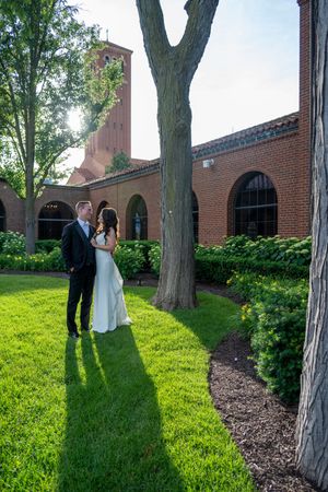 Bride and groom stand together on lush green lawn beside brick church building with arched windows and tower.