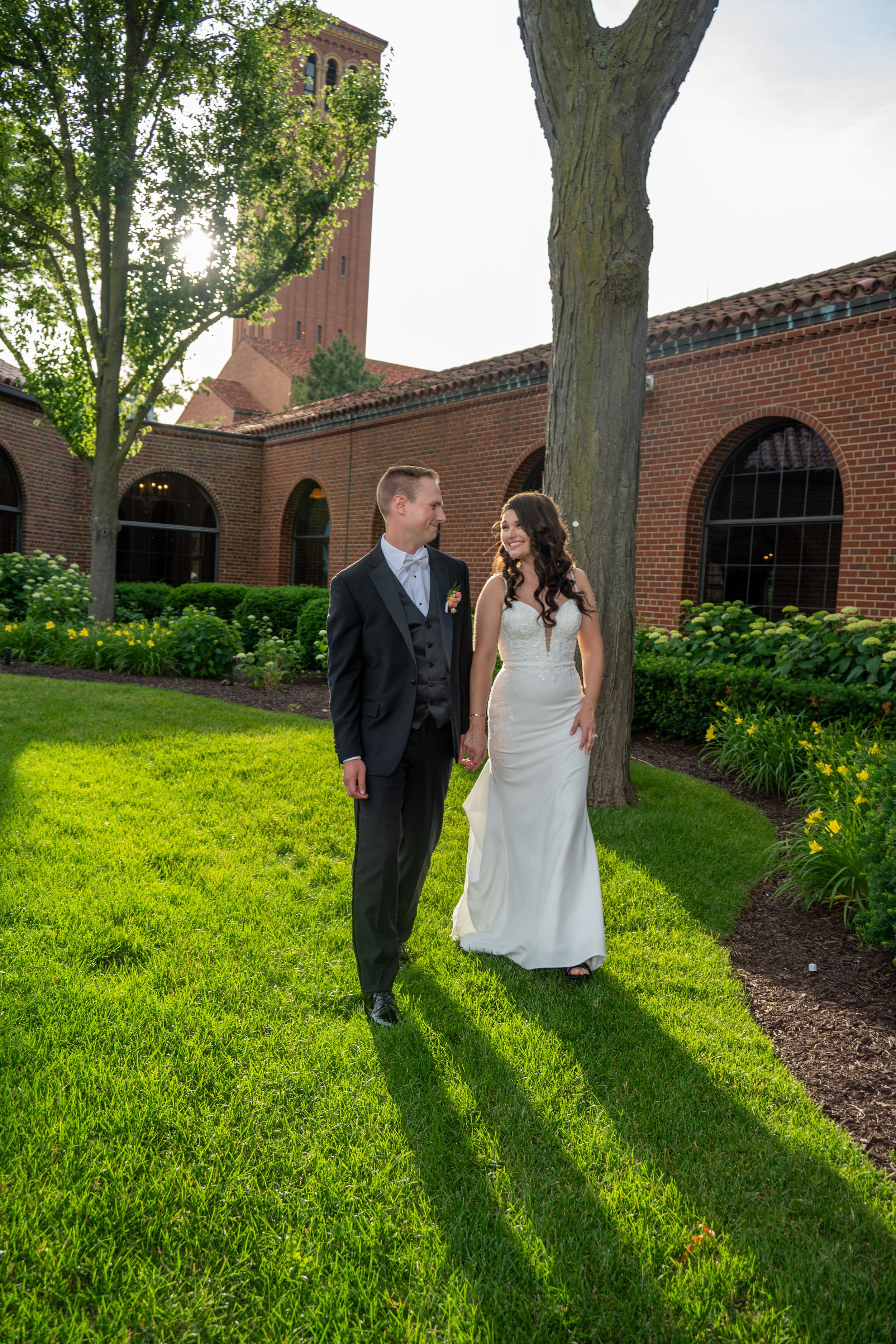 A bride and groom walk hand in hand on lush green grass beside a historic brick building on their wedding day.