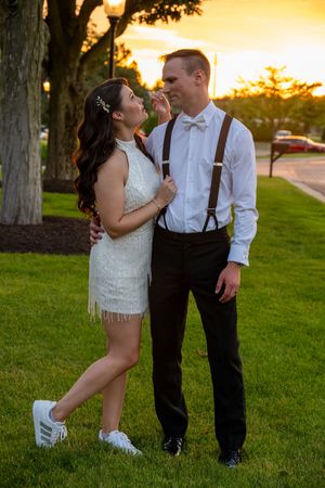 Couple in wedding attire poses at sunset on green lawn, woman in white fringe dress, man in suspenders and bow tie.