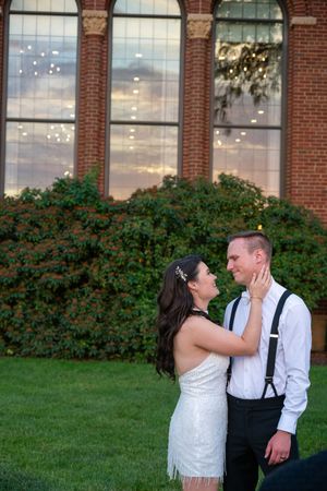 Bride and groom share a romantic moment on green lawn before brick building with large windows at golden hour.