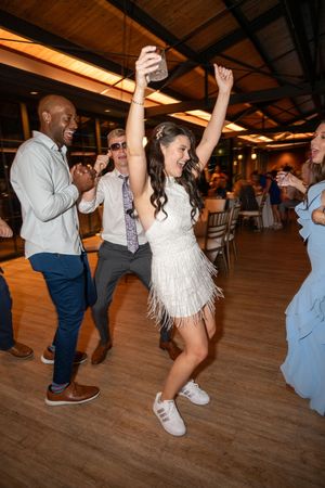 Bride in white fringe dress and sneakers dancing joyfully with guests on wedding reception dance floor.
