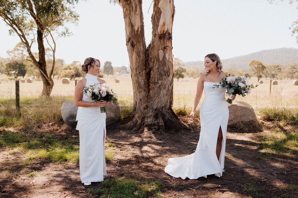 Two brides in white gowns holding bouquets stand near a eucalyptus tree in a rural Australian landscape.