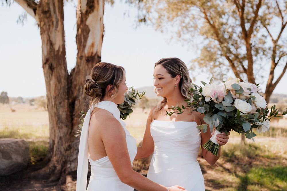 Two brides in white gowns share a joyful moment holding a bouquet of white roses and greenery outdoors.