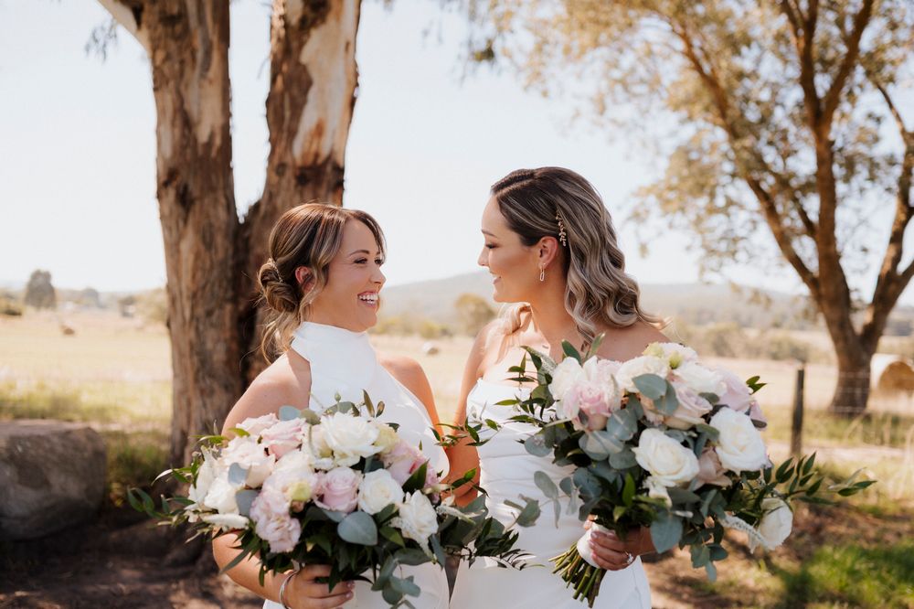 Two brides in white gowns holding white floral bouquets smiling at each other outdoors on a sunny wedding day.