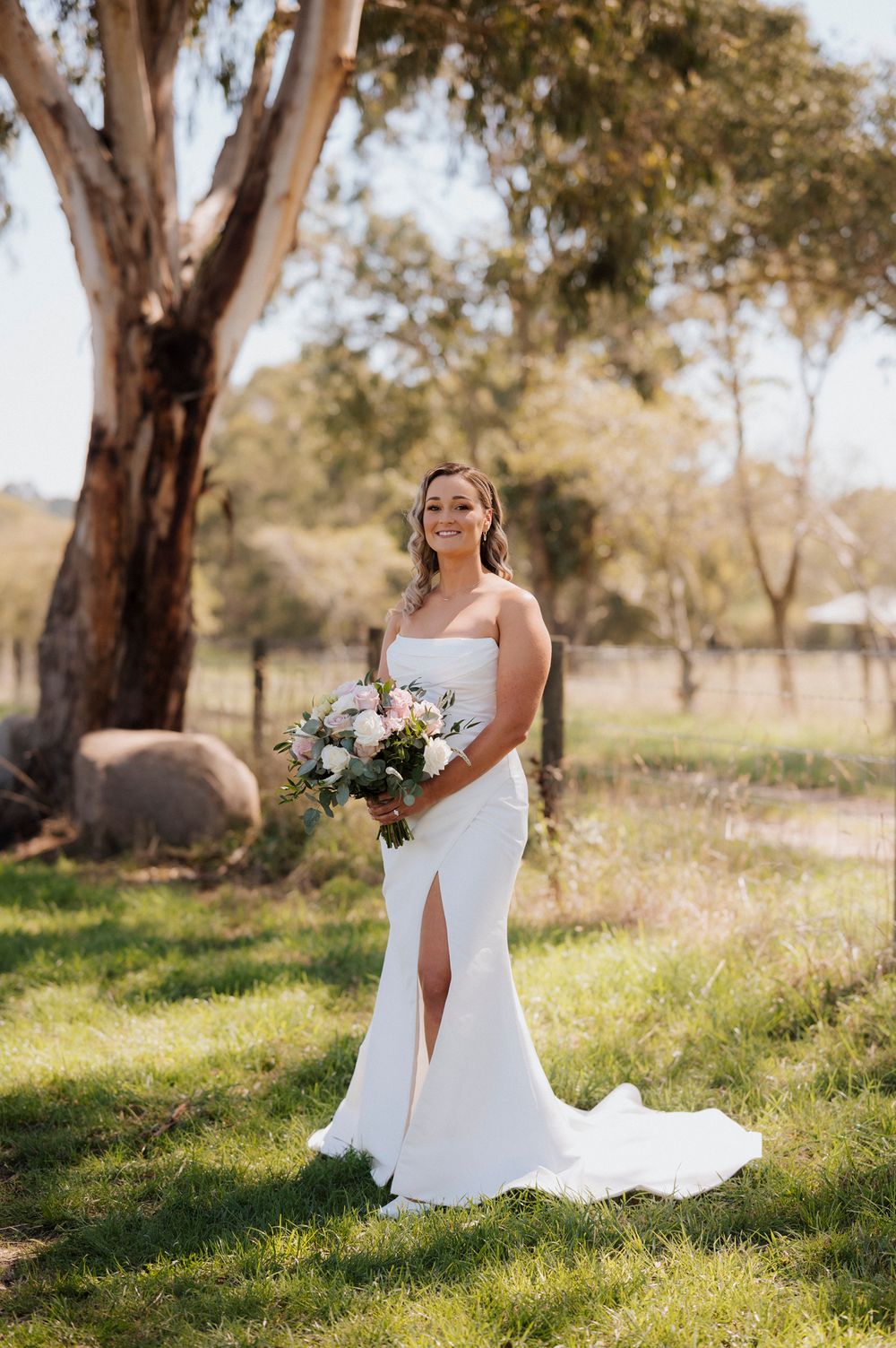 Bride in white strapless gown with slit holds bouquet outdoors near eucalyptus tree on green lawn.