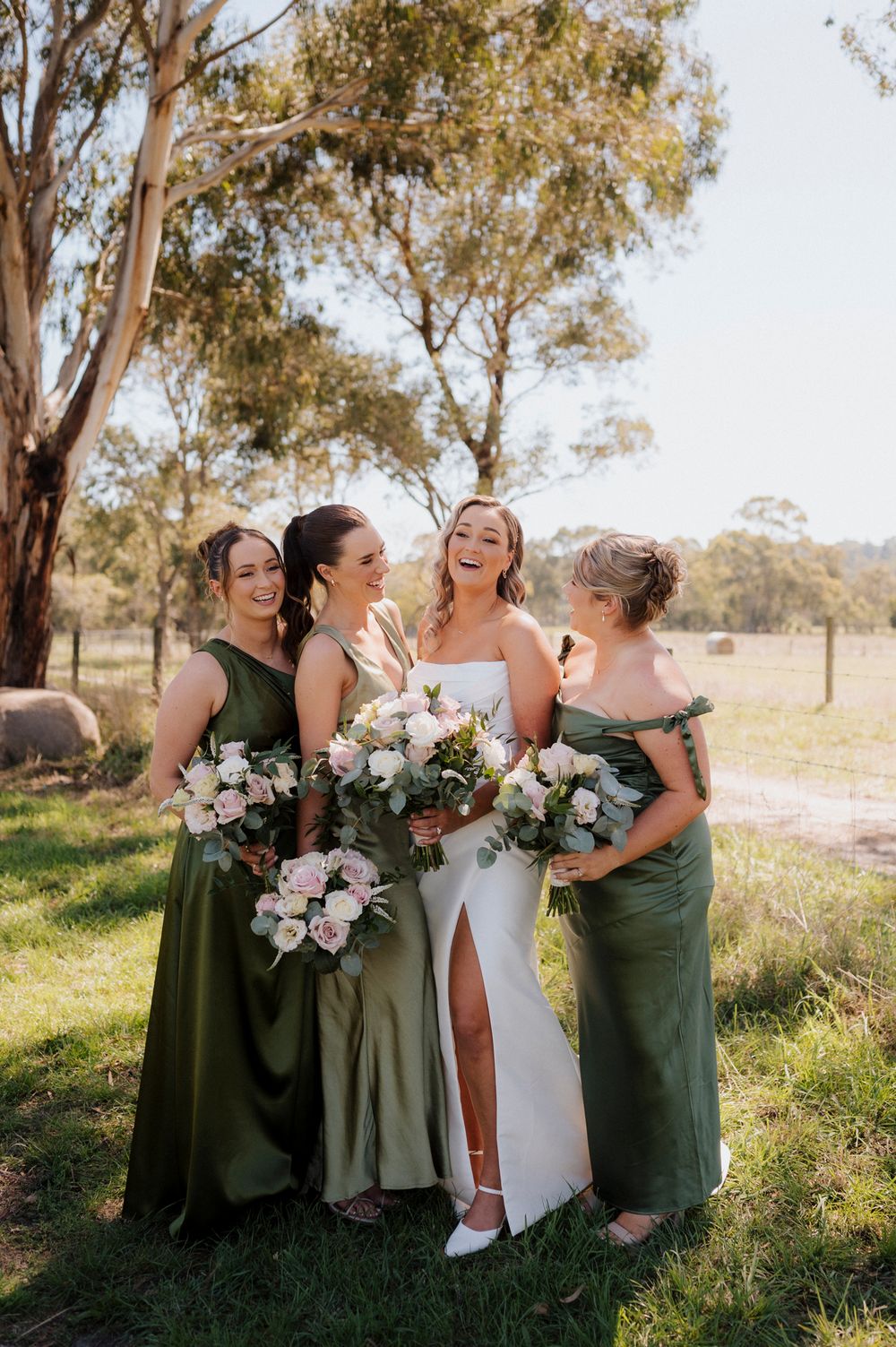 Bride in white gown laughing with three bridesmaids in olive green dresses holding pink bouquets outdoors.