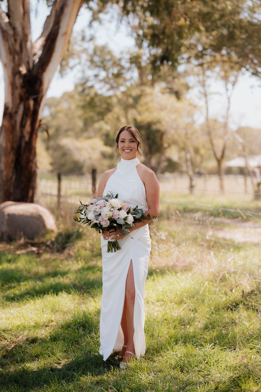 Bride in white halter neck gown with front slit holds pink bouquet, standing on green grass with eucalyptus trees.
