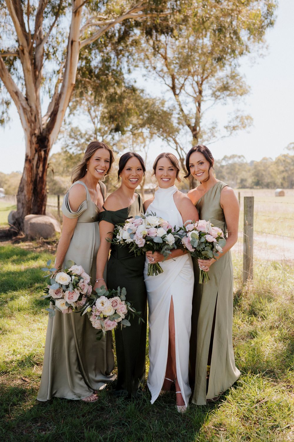 Bride in white gown with three bridesmaids in sage green dresses holding pink bouquets outdoors.