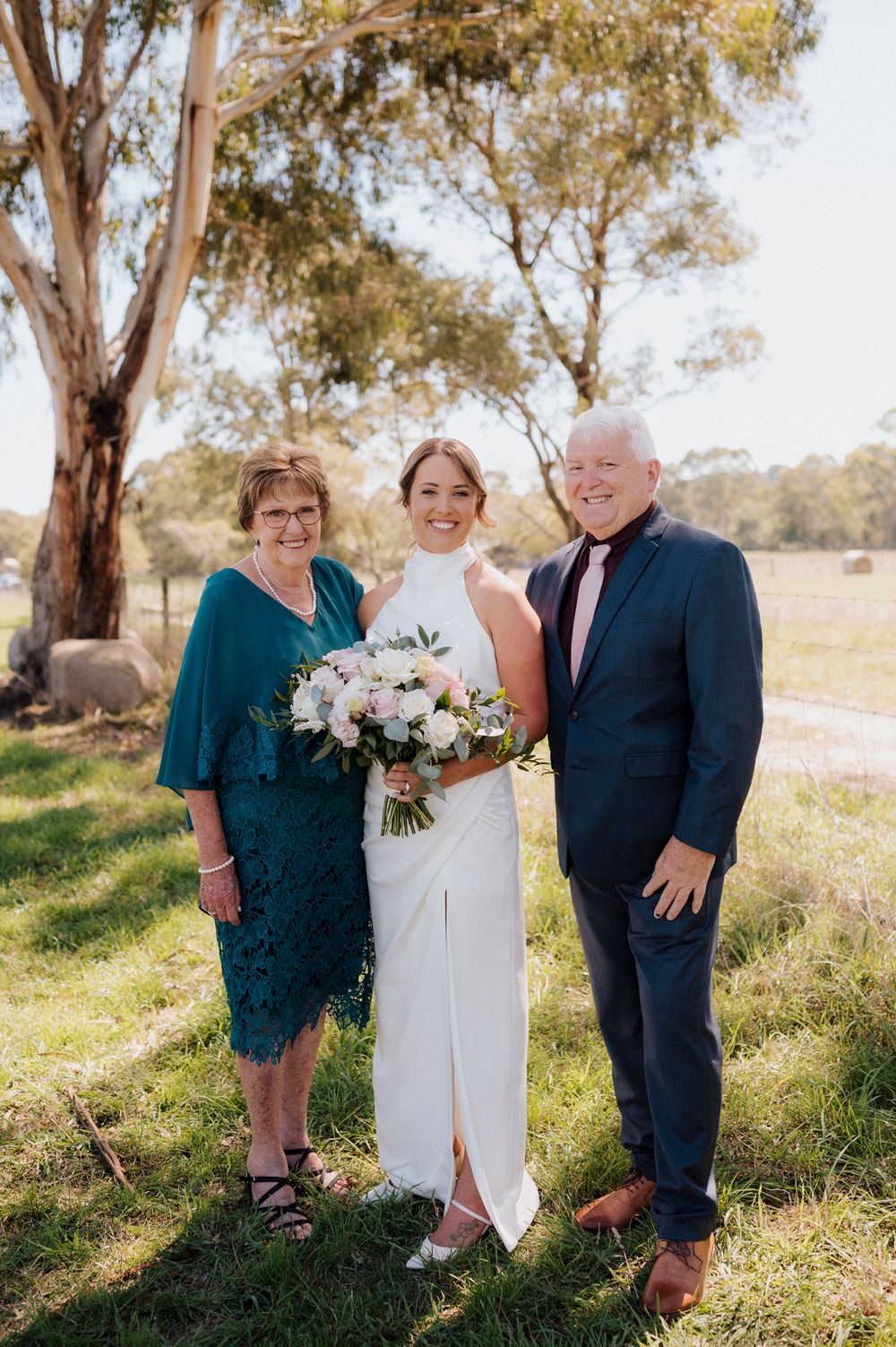 Bride in white halter gown holds bouquet flanked by parents in formal attire at outdoor rural wedding venue.