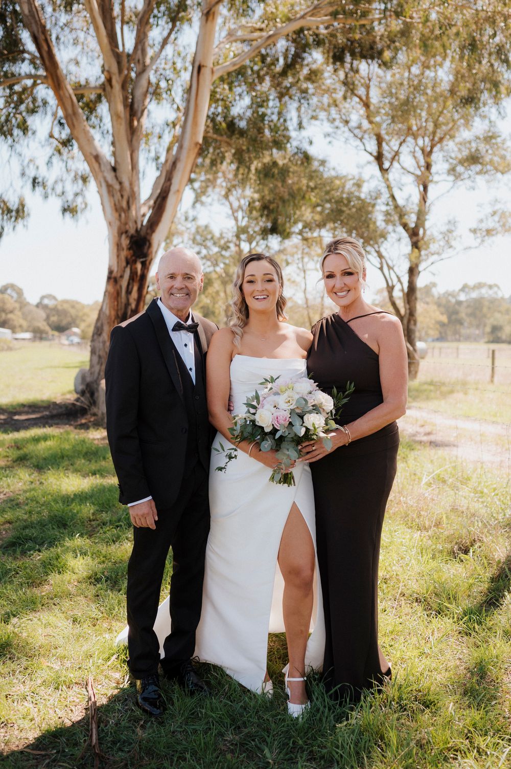 Bride in white gown with bouquet poses with man in tuxedo and woman in black dress outdoors.