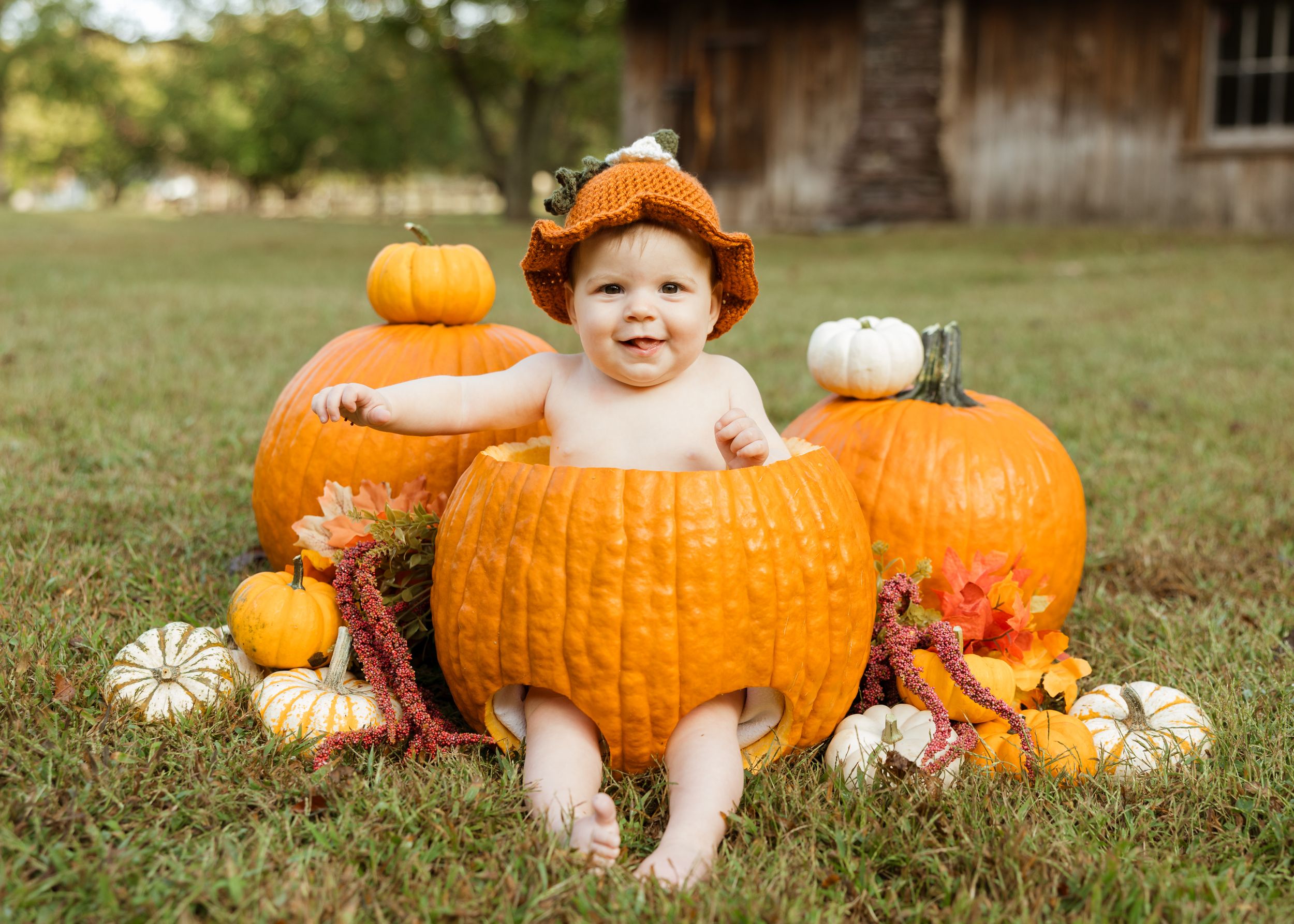 Smiling baby sitting inside a large pumpkin surrounded by colorful gourds outdoors in fall.