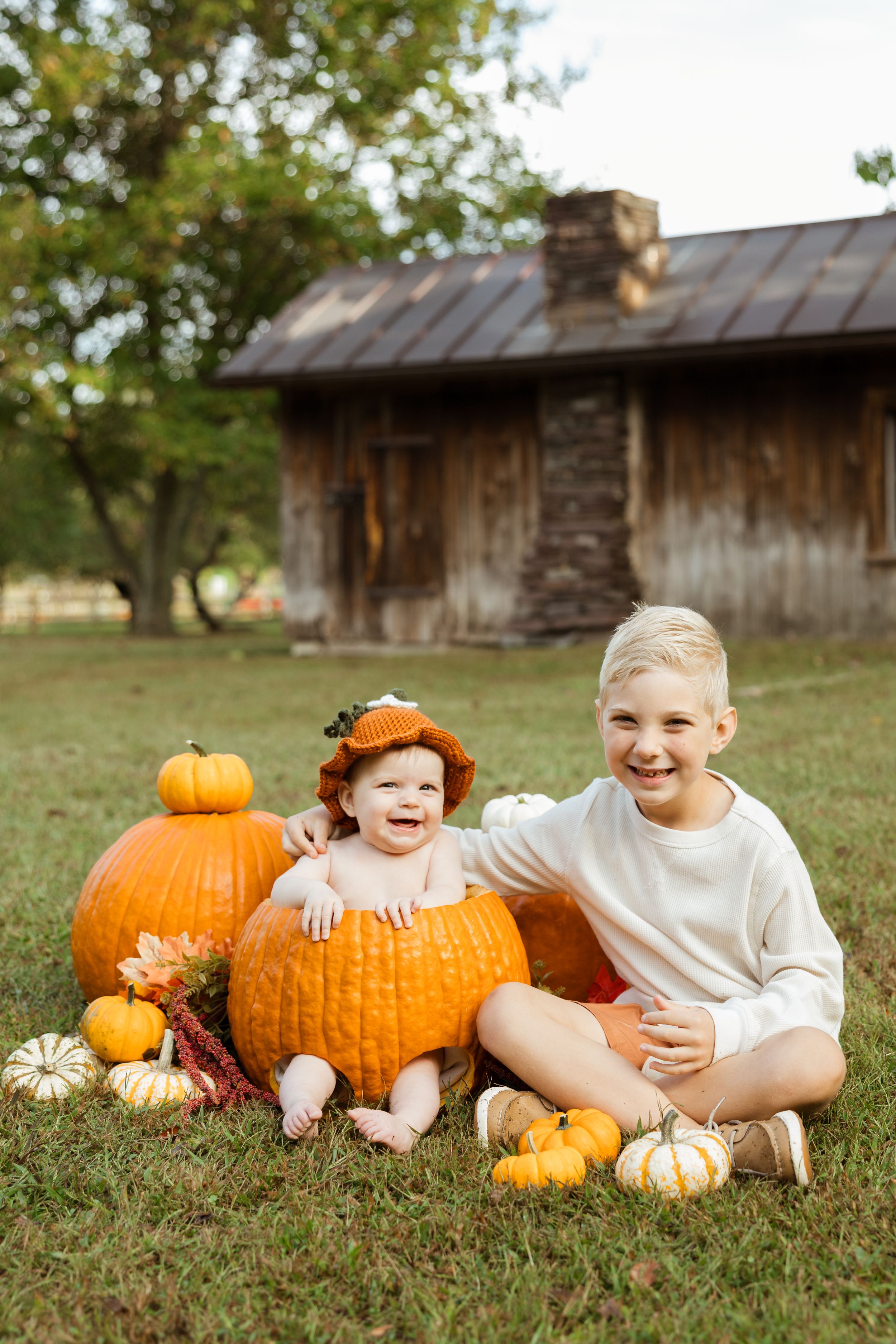 Baby in pumpkin with older sibling laughing outdoors, fall harvest scene with pumpkins and rustic barn background.
