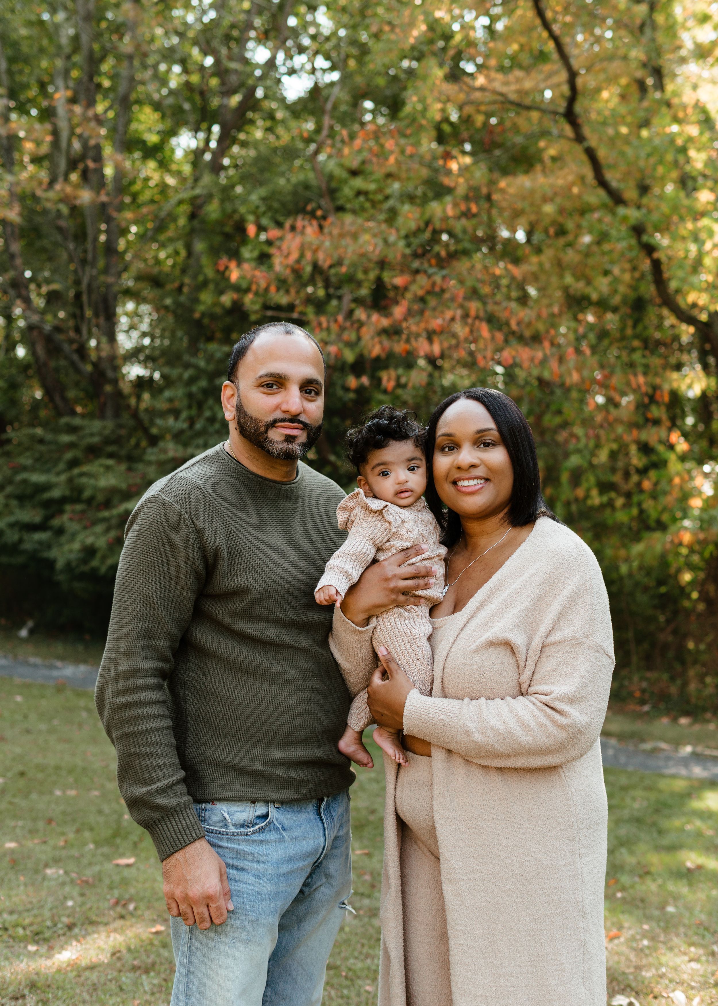 A family of three poses outdoors in fall, parents holding their baby, surrounded by colorful autumn foliage.
