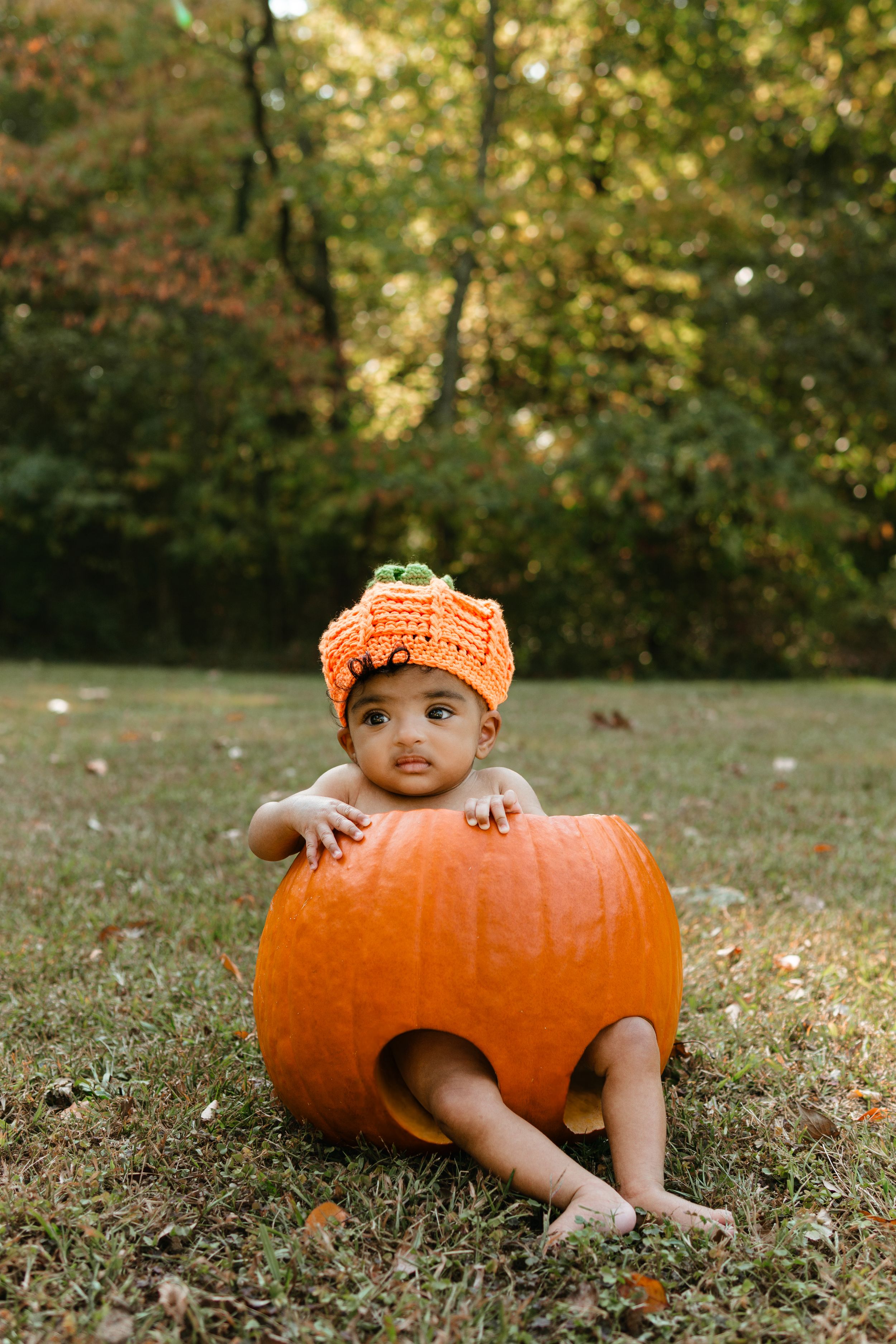 Adorable baby in orange crochet pumpkin hat sitting inside a large pumpkin on grass with autumn trees in background.