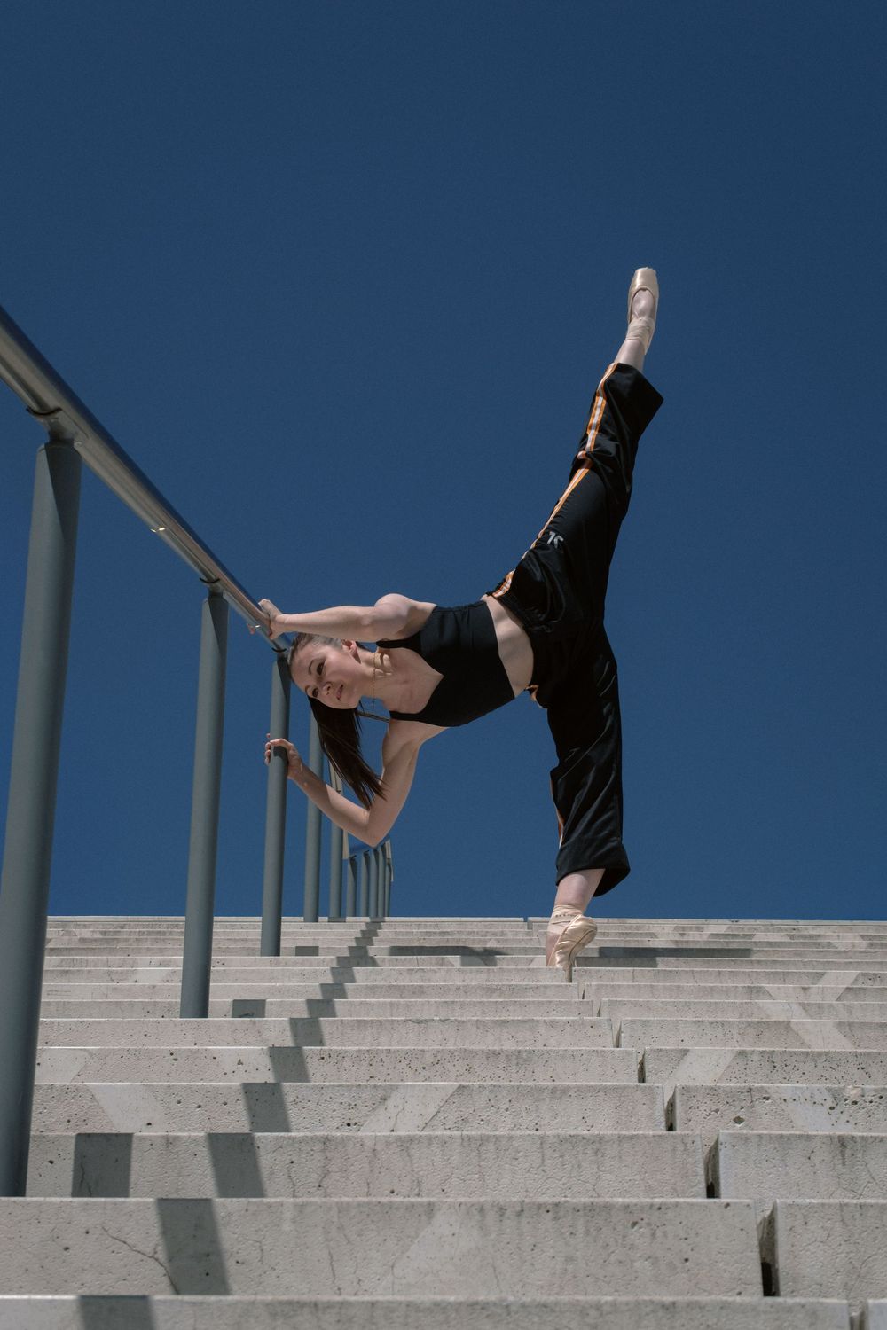 Danseuse classique en équilibre sur un escalier en béton sous un ciel bleu.