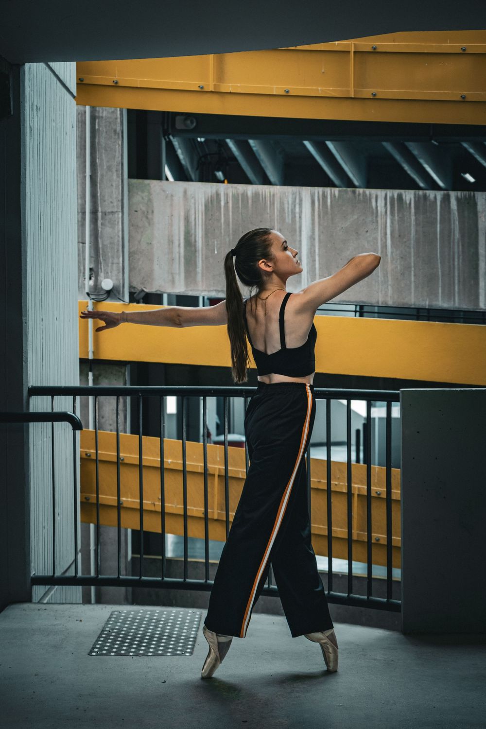 Danseuse classique en mouvement dans un parking aux structures de béton et de métal jaune.