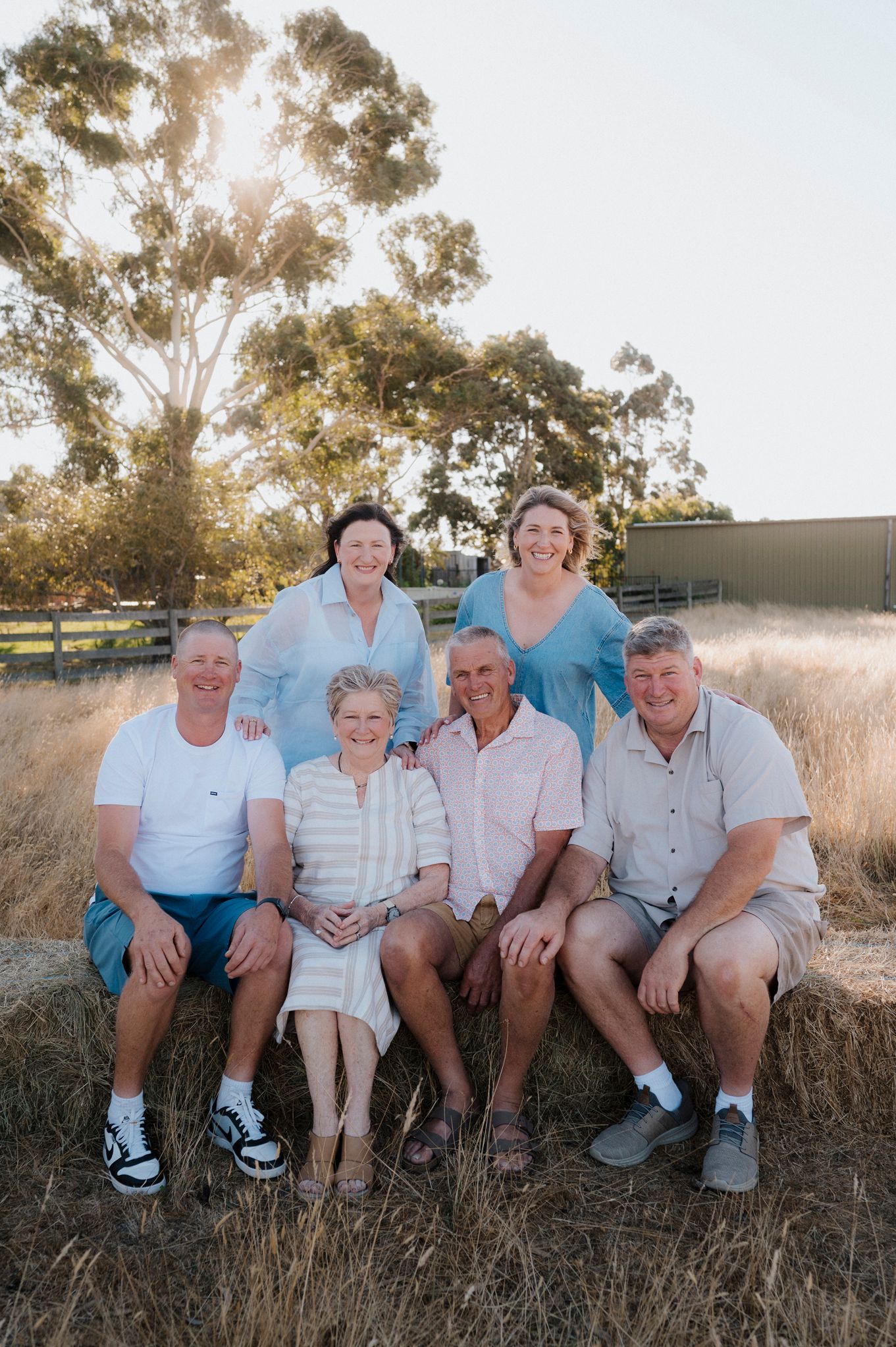 Six family members pose together on hay bales in a rural Australian farm setting at golden hour.