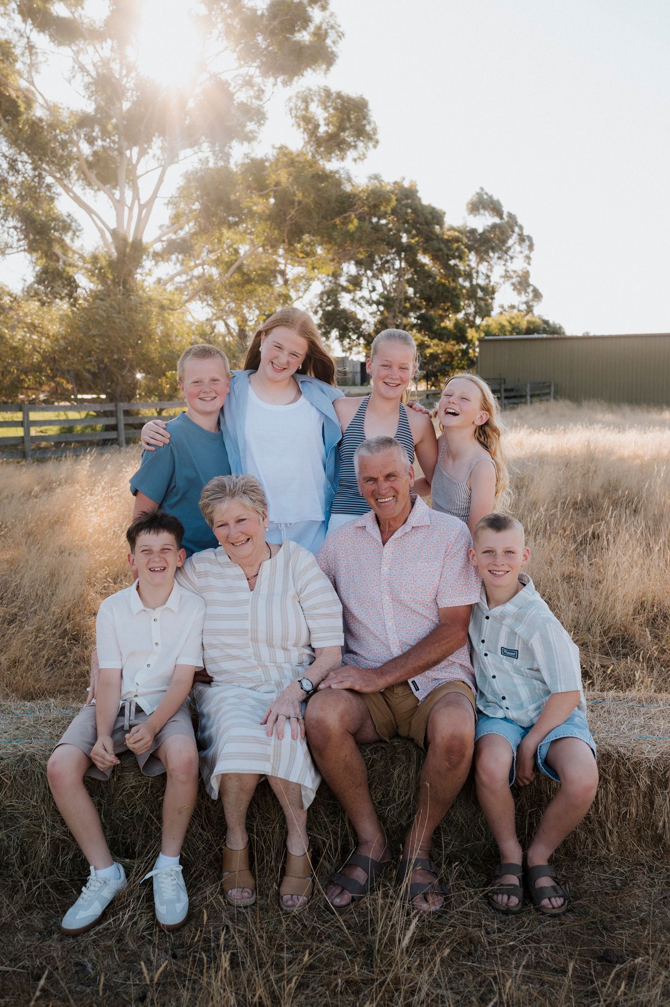 Grandparents seated on hay bale surrounded by four grandchildren in sunny rural farm setting at golden hour.
