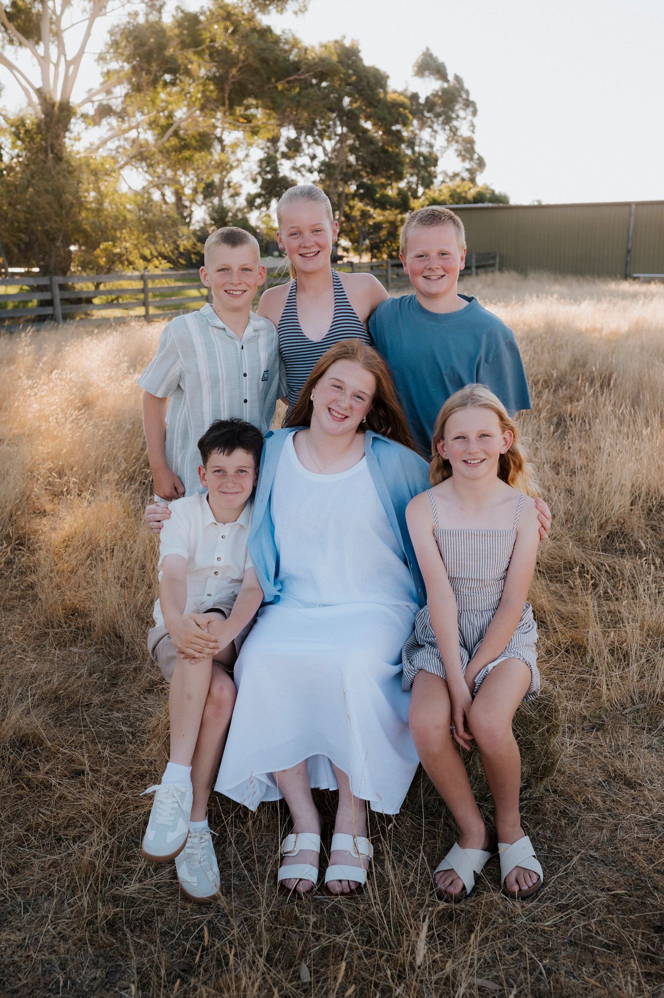 Family of six posing outdoors in golden field at sunset, with children smiling warmly in casual summer attire.