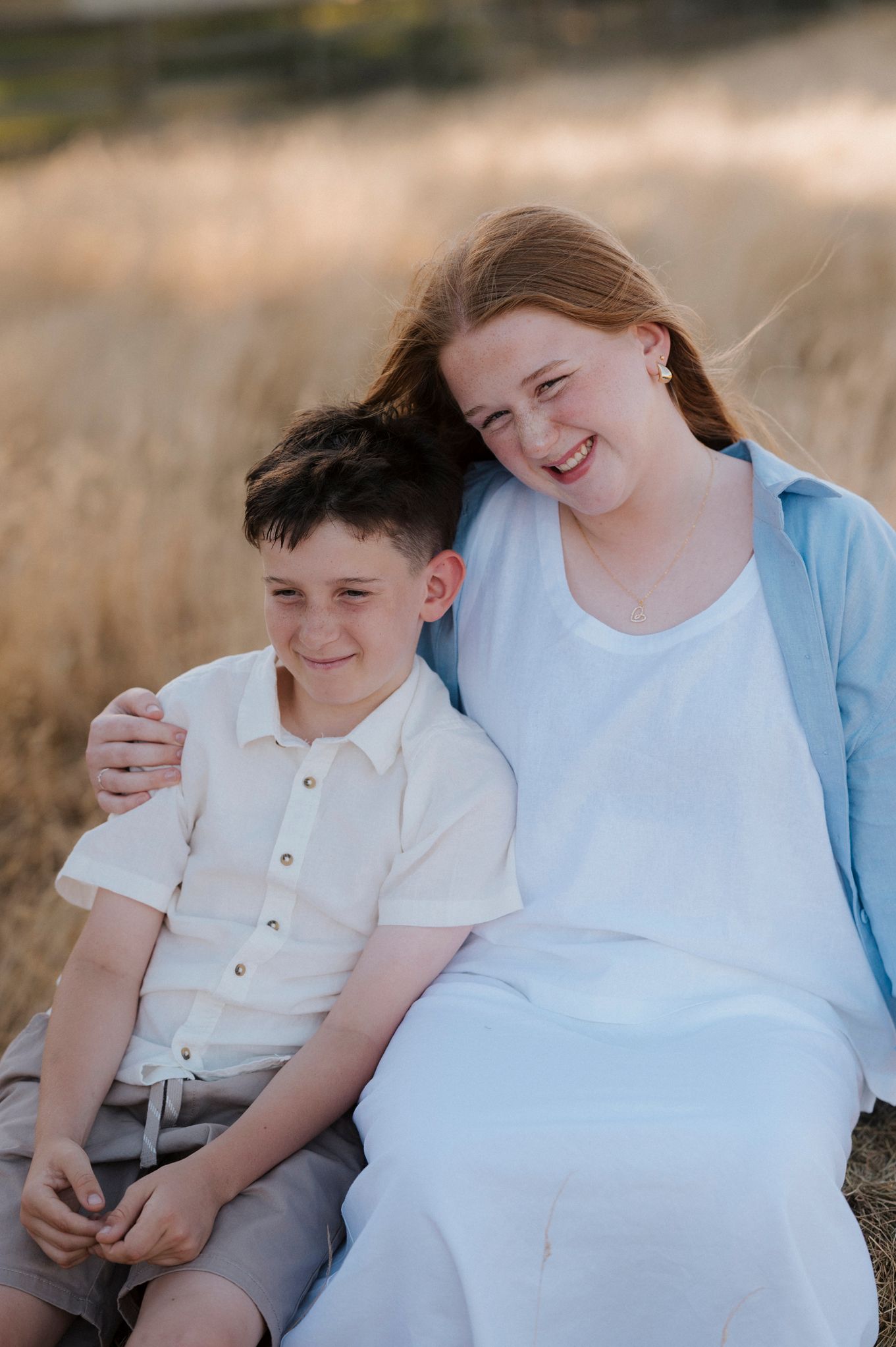 A teenage girl with red hair hugs a young boy while sitting together in a golden field, both smiling warmly.