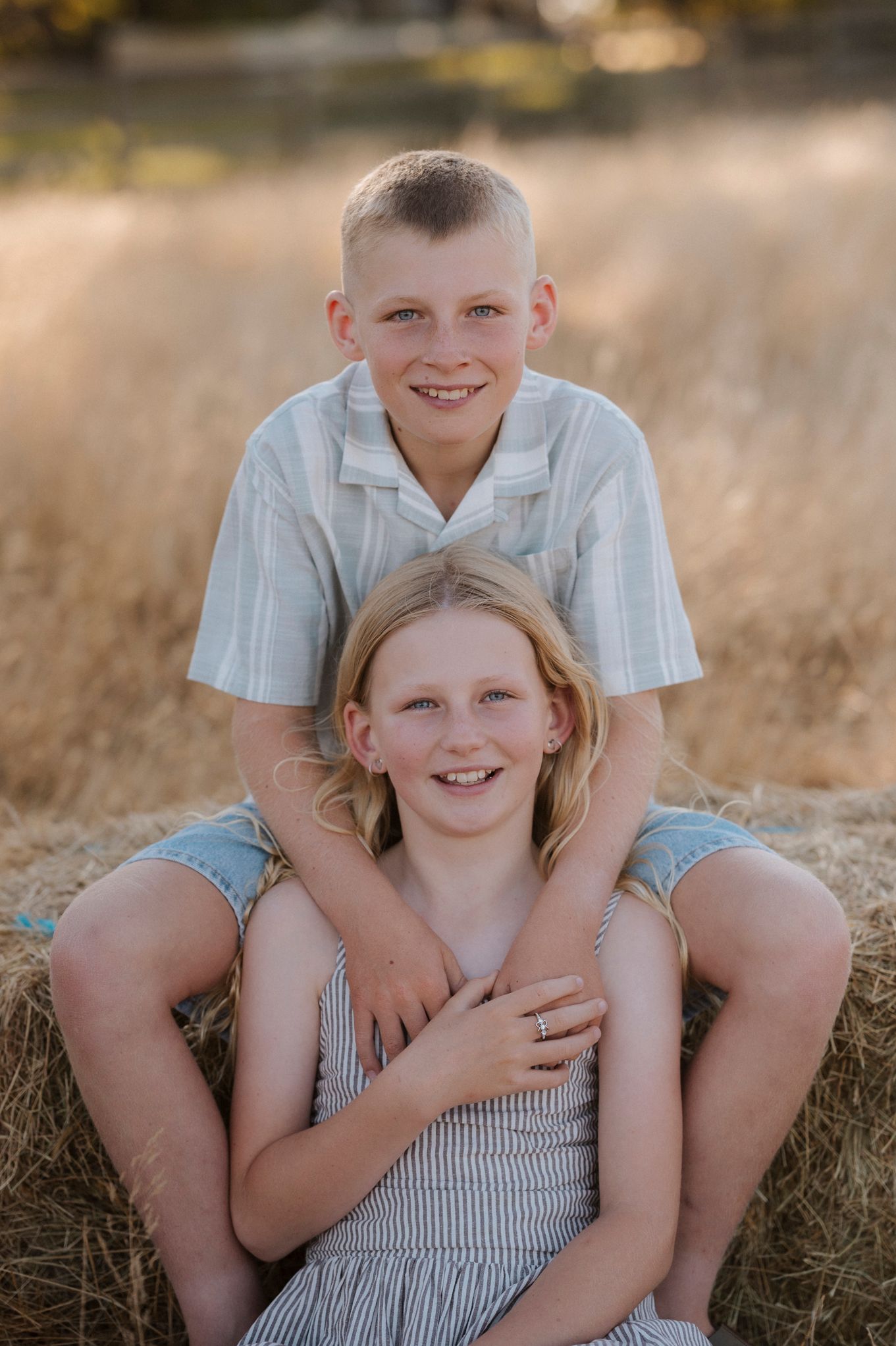 Two smiling children pose on a hay bale in a golden field, boy sitting behind girl in a warm outdoor portrait.