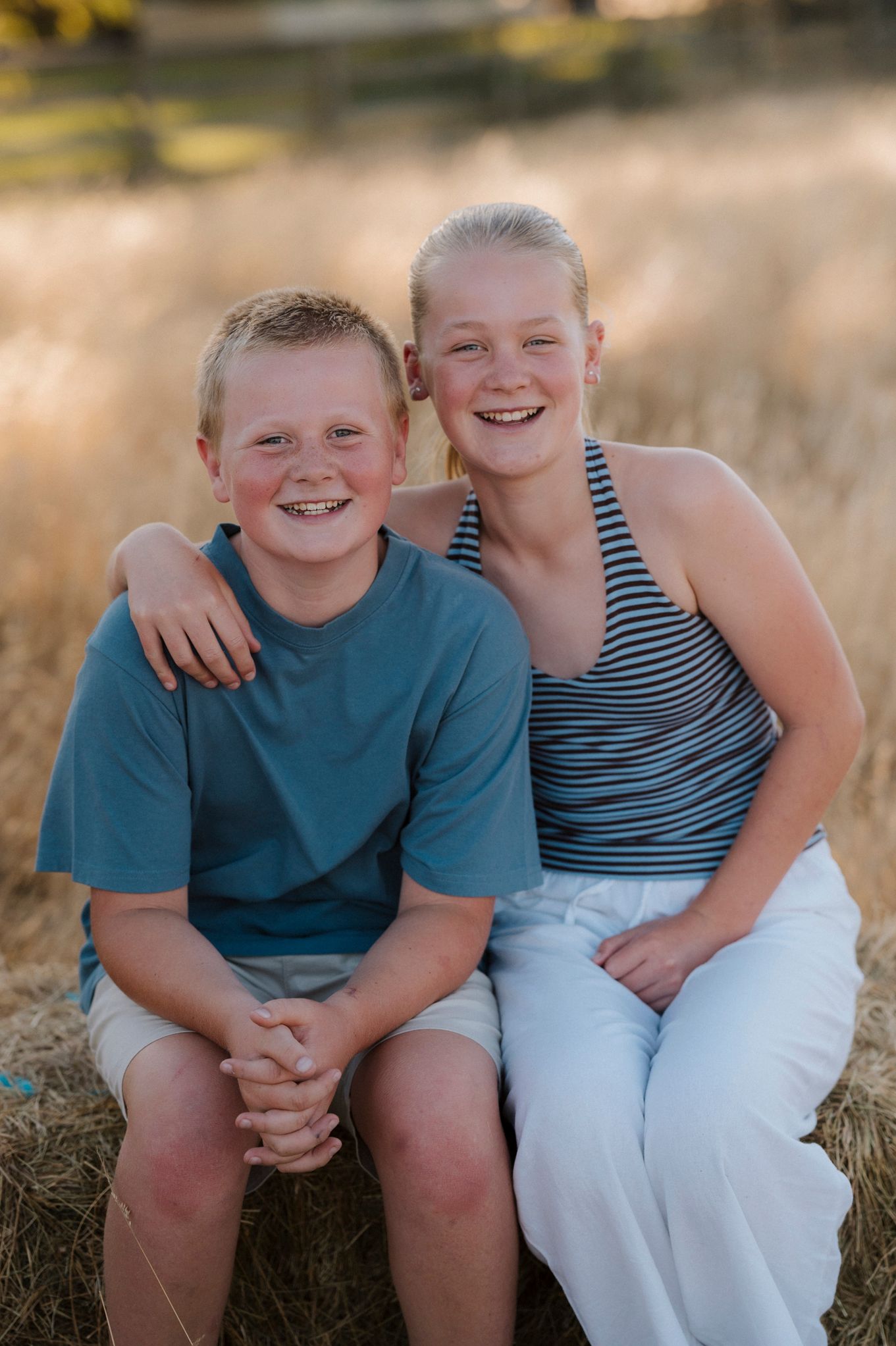 Two smiling children sit on a hay bale in a golden field, boy in blue tee and girl in striped top with white pants.