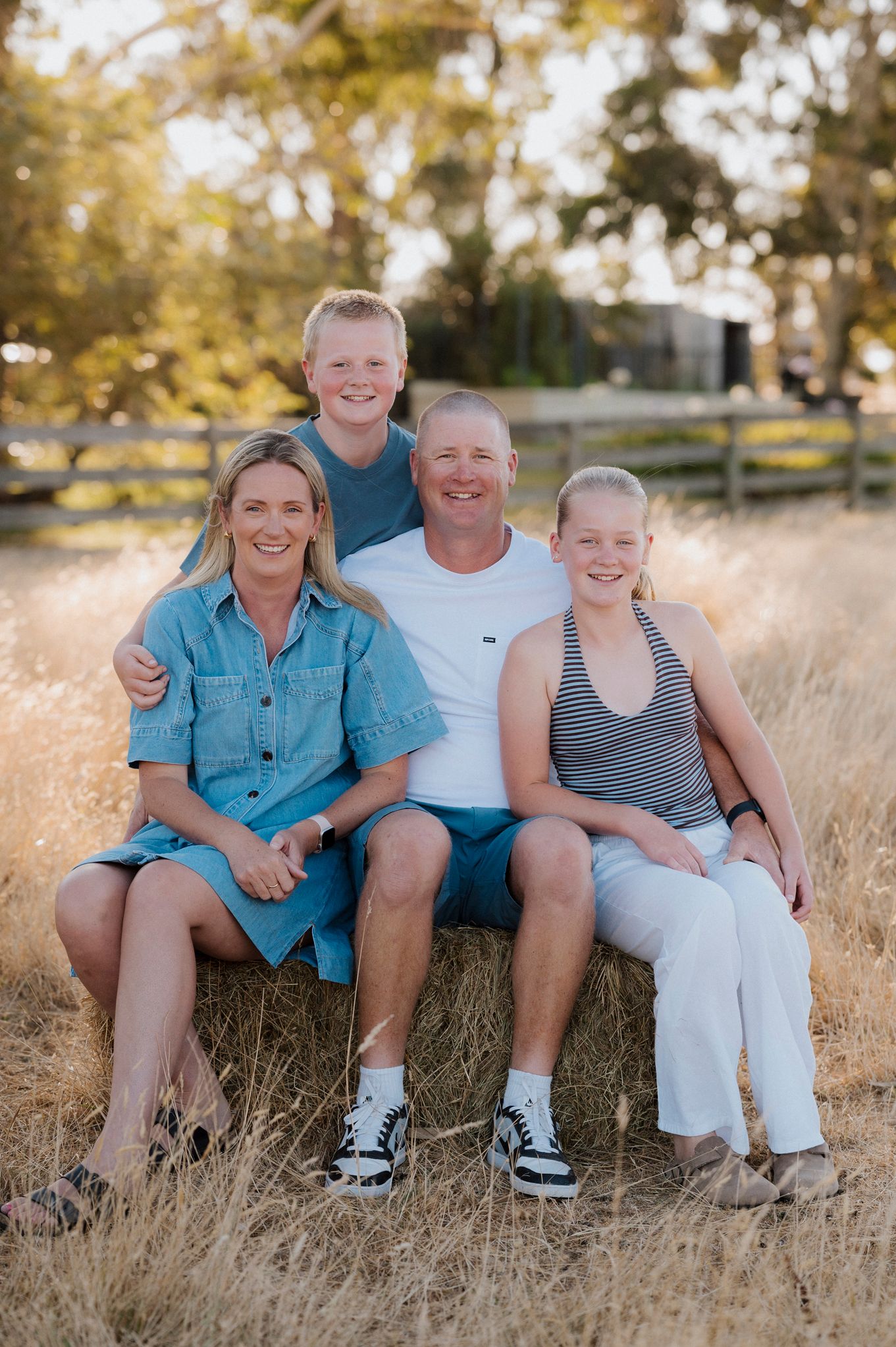 Family of four smiling on a hay bale in a golden field during a sunny outdoor photo session.