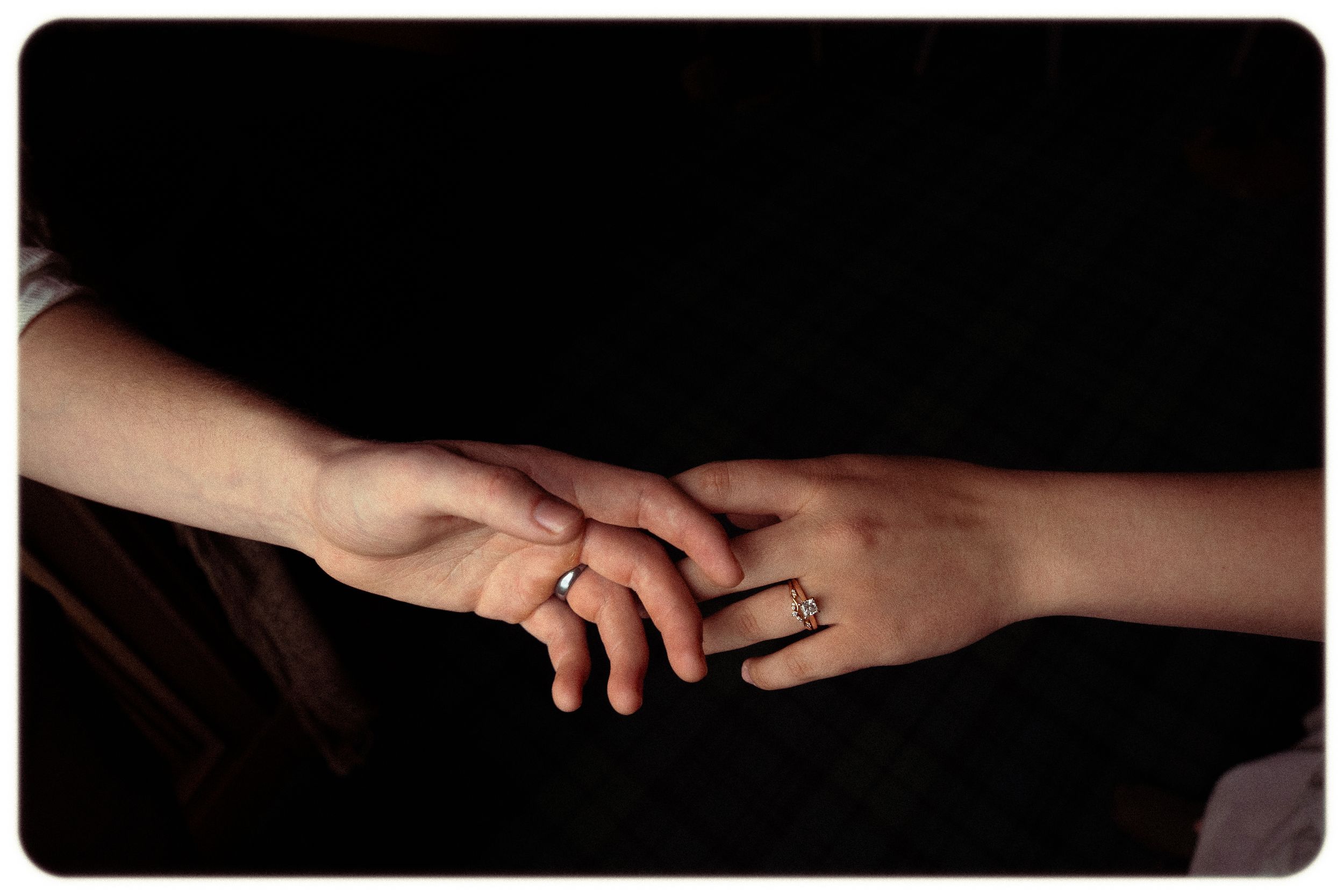 Two hands gently touching, both wearing rings, against a dark background, symbolizing love and connection.