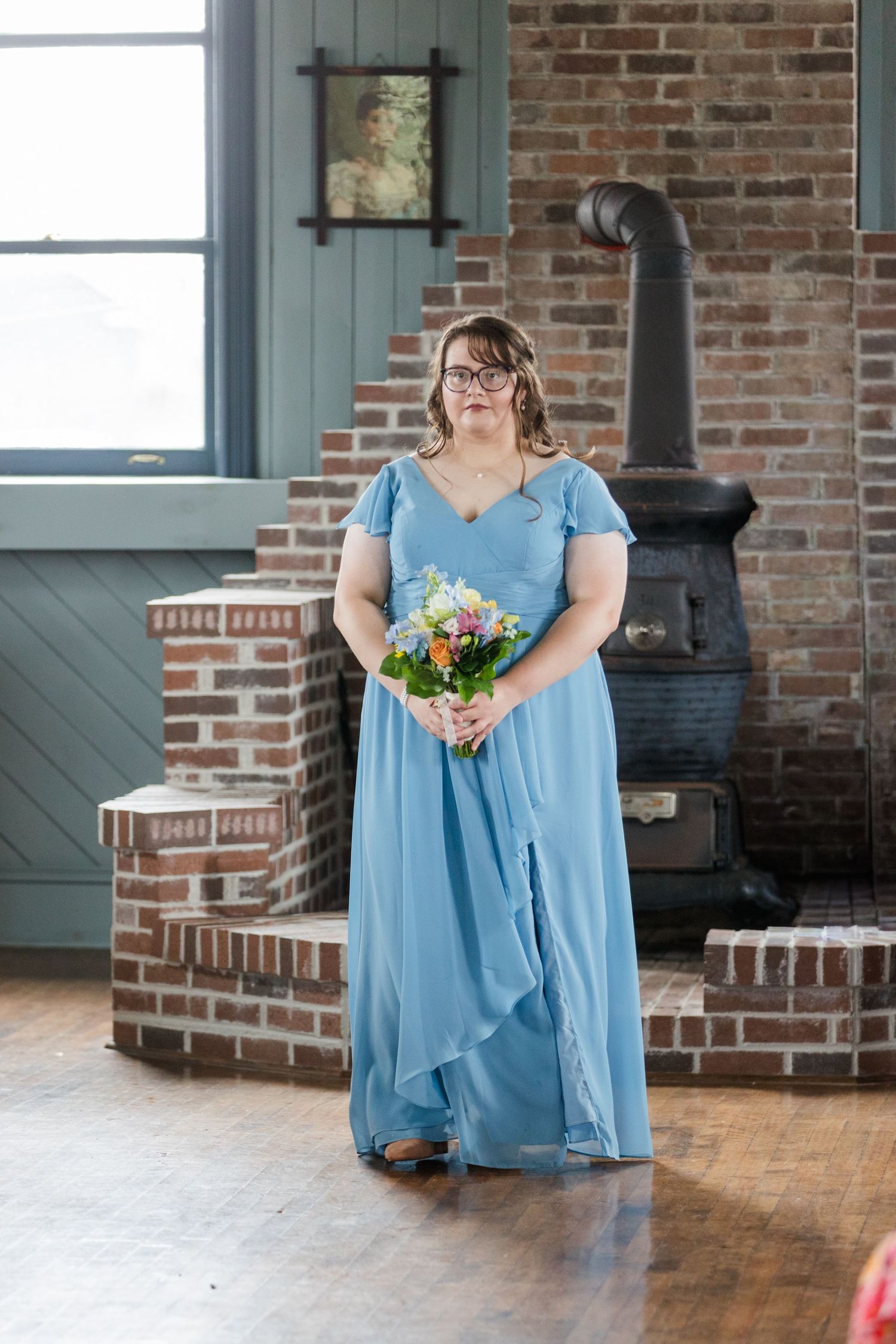 Bridesmaid in blue gown holding floral bouquet stands near brick fireplace with wood stove in rustic venue.