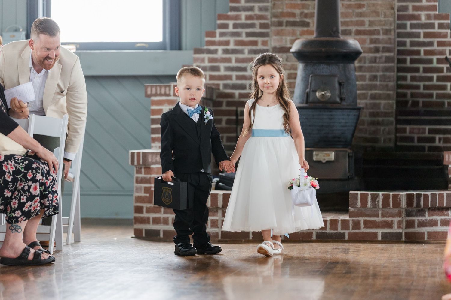 Young ring bearer and flower girl in formal attire hold hands at a wedding ceremony near a brick fireplace.