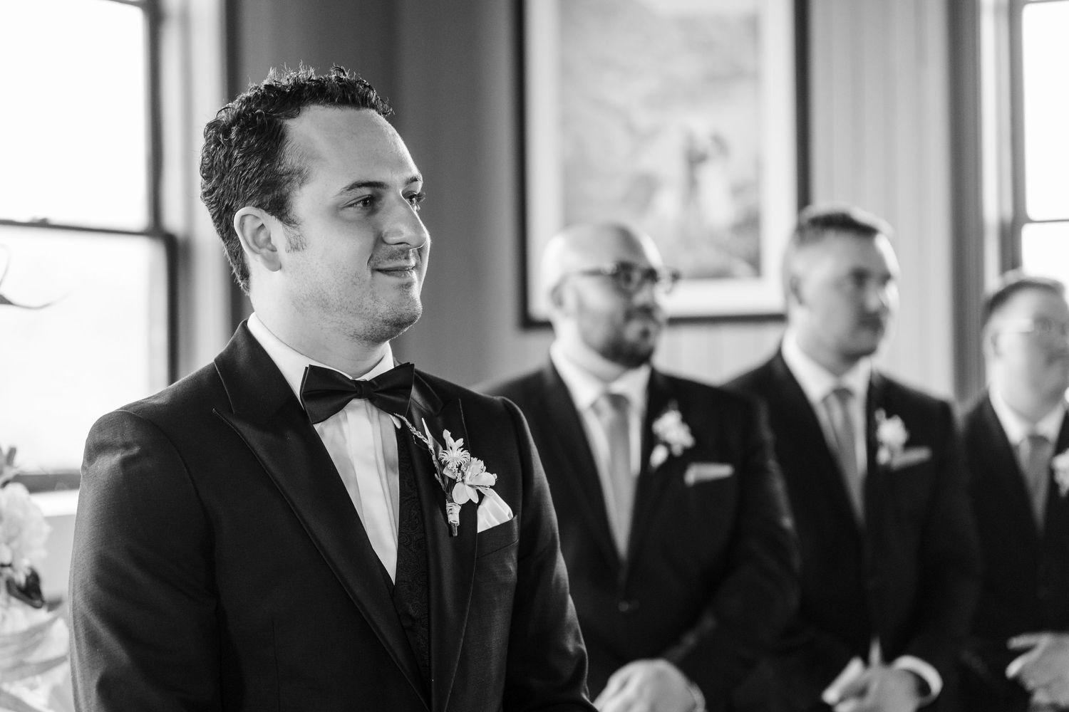 Groom smiling at altar in black tuxedo with boutonniere, groomsmen standing behind in black and white wedding photo.