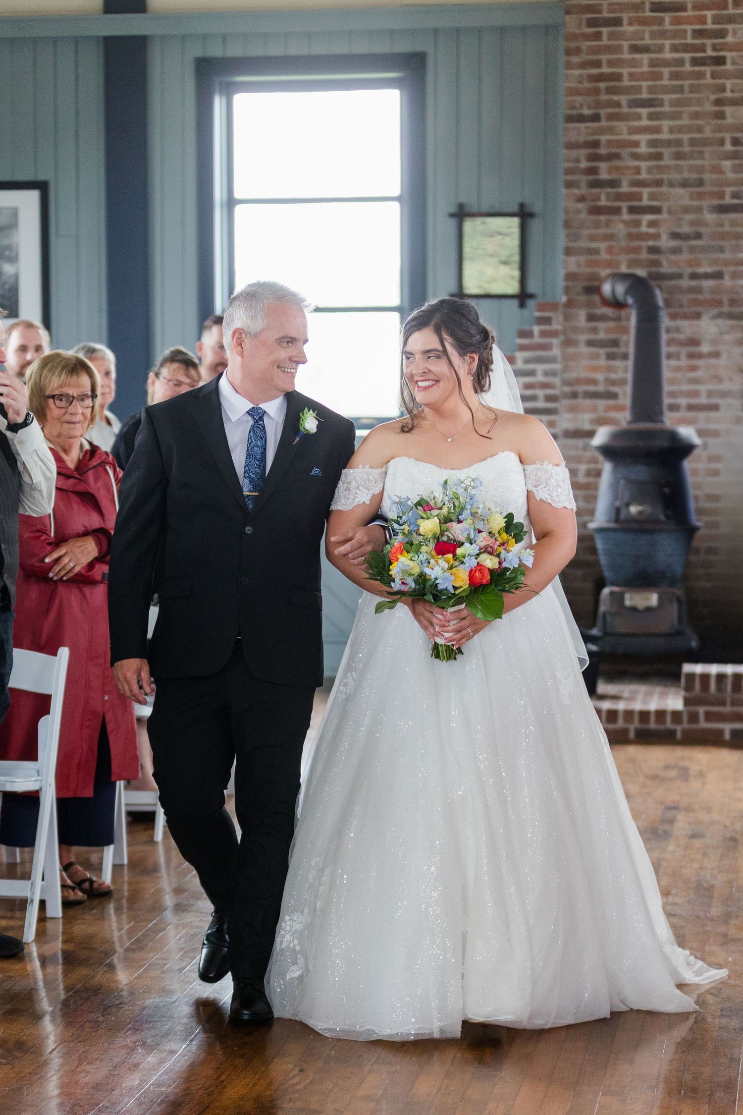 Bride in white off-shoulder gown walks down aisle with father, holding colorful bouquet at indoor wedding ceremony.