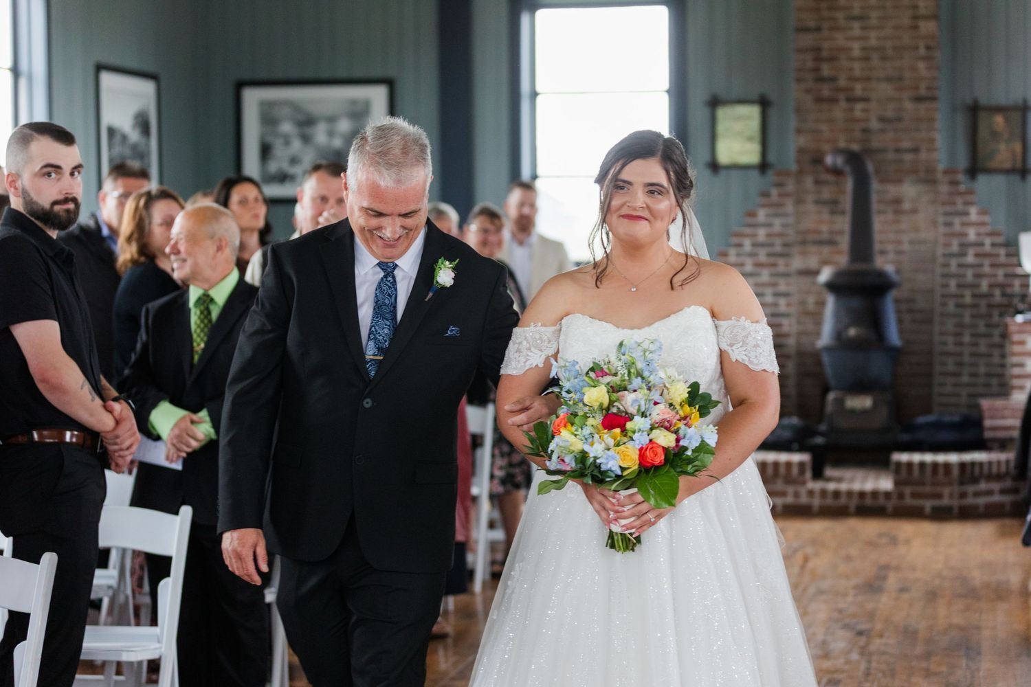 Bride in white off-shoulder gown walks down aisle with escort holding colorful bouquet at indoor wedding ceremony.