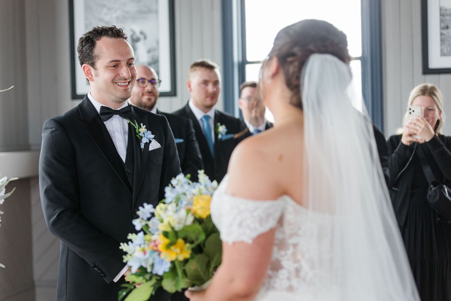 Groom in black tuxedo smiling at bride in white lace dress and veil during indoor wedding ceremony.