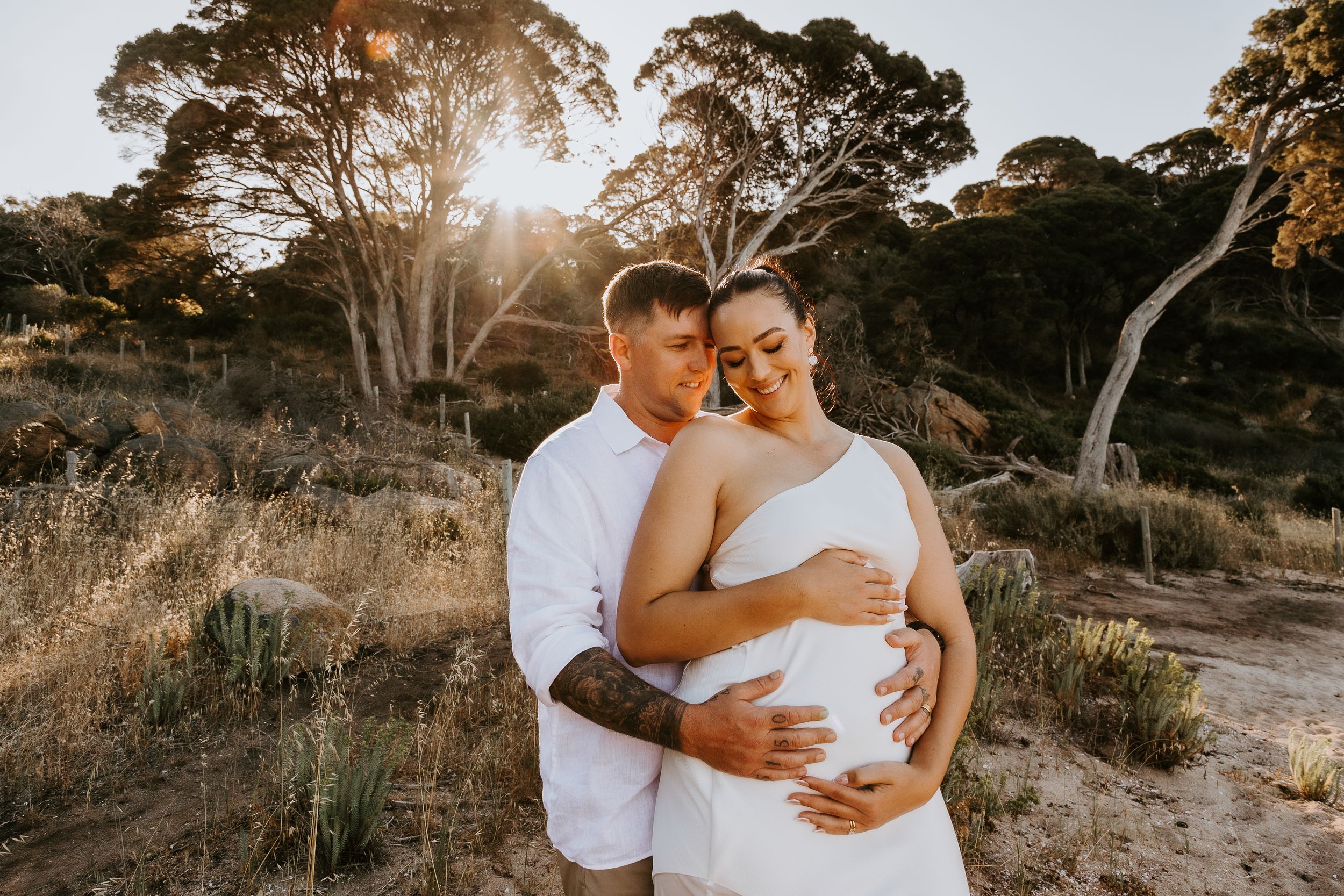 Couple embracing outdoors at golden hour, pregnant woman in white dress with partner holding her belly among eucalyptus trees.