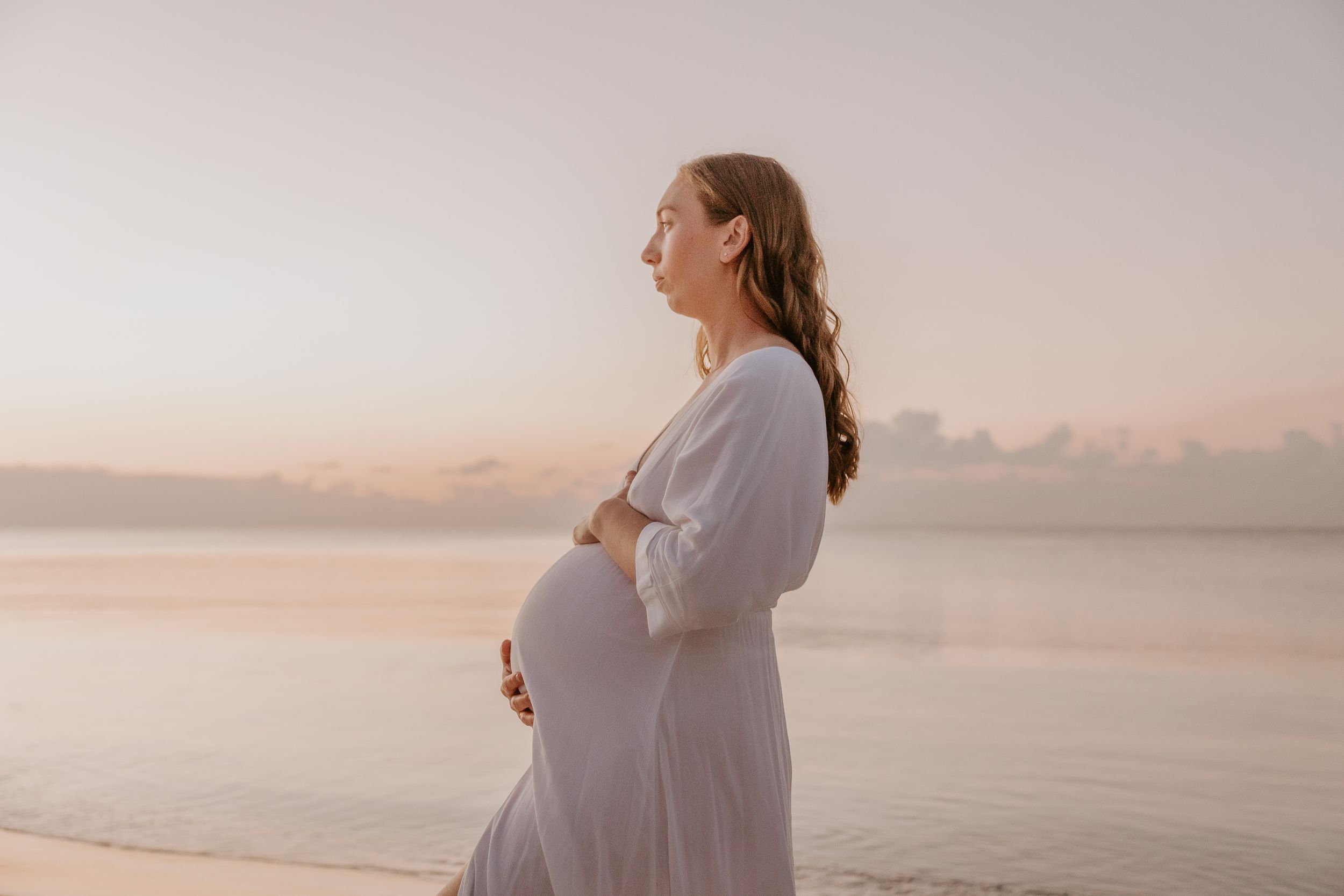 Pregnant woman in white dress cradling her belly on a serene beach at sunset.