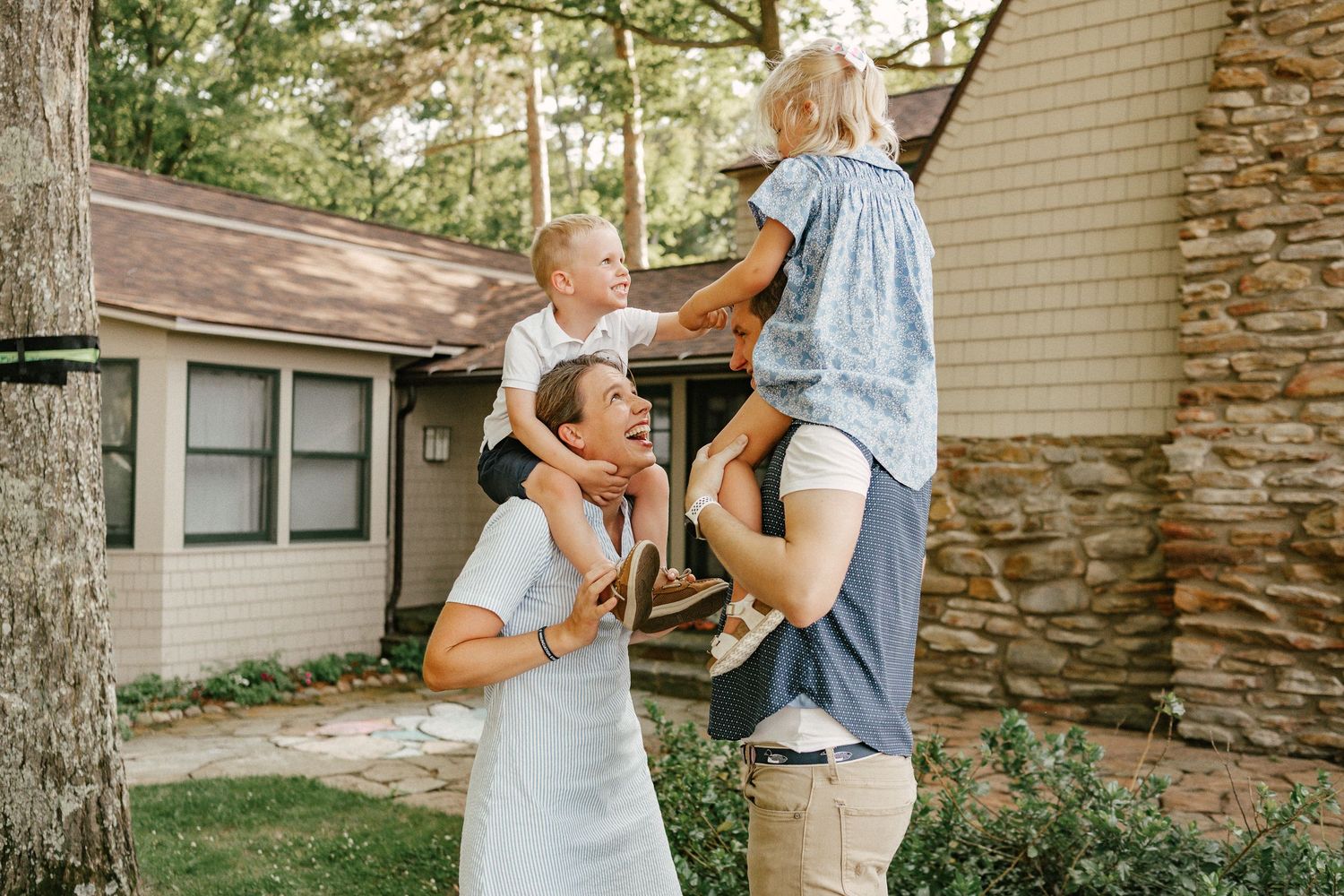Family of four enjoying time outside a rustic cabin, with children on parents shoulders, surrounded by trees.